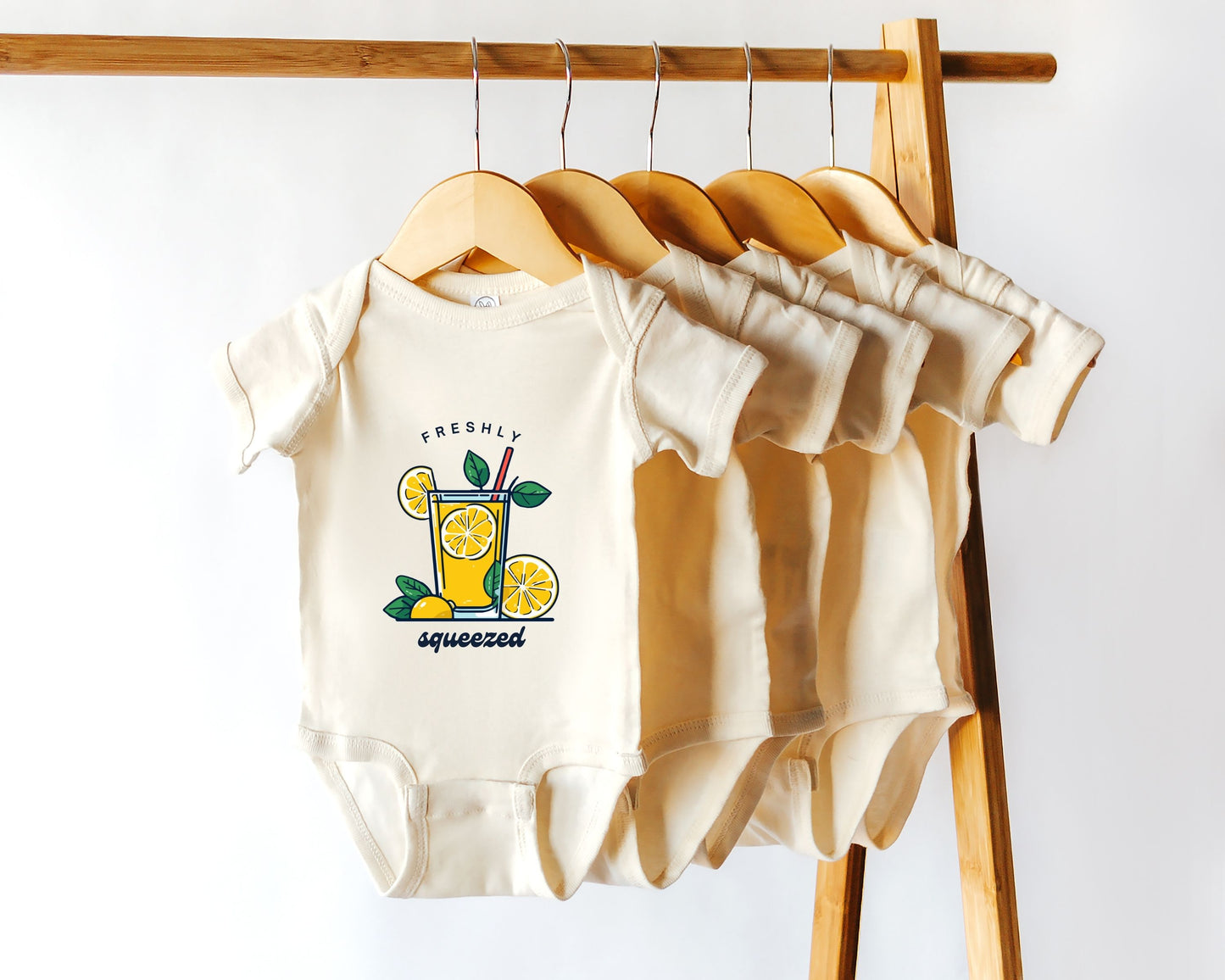 A short sleeve cotton baby bodysuit with a lemonade graphic and the text 'Freshly Squeezed Lemonade' hanging on a wooden rack among other similar bodysuits.