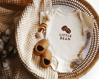 A short sleeve coffee themed baby onesie with a coffee bean graphic and the text little bean displayed in a wicker basket.