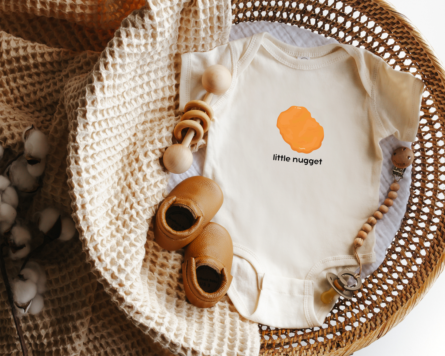 A baby bodysuit laid flat in a woven basket with styling props, including a cream knit blanket. The bodysuit is off-white with a chicken nugget graphic and the text 'little nugget' on the chest.