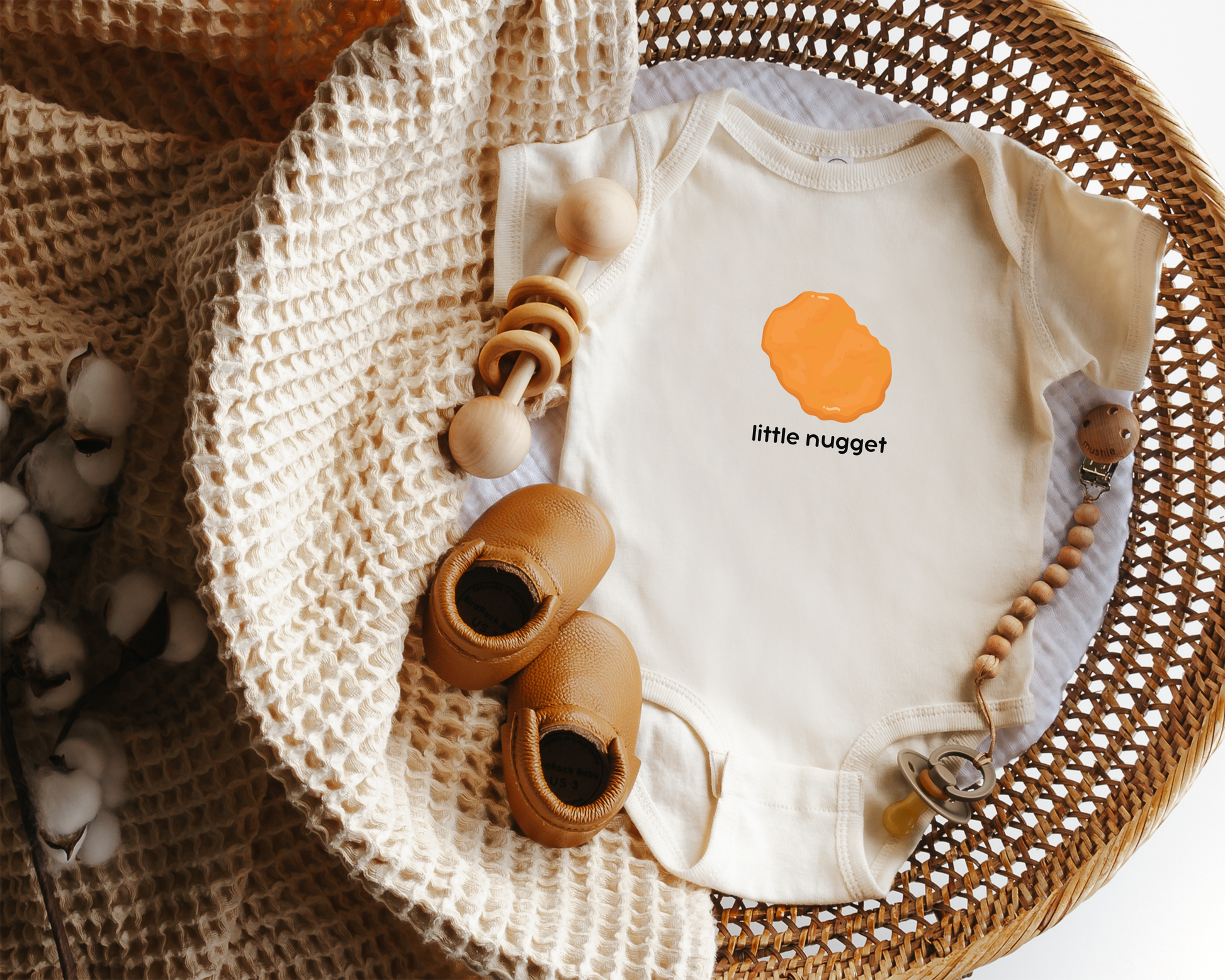 A baby bodysuit laid flat in a woven basket with styling props, including a cream knit blanket. The bodysuit is off-white with a chicken nugget graphic and the text 'little nugget' on the chest.