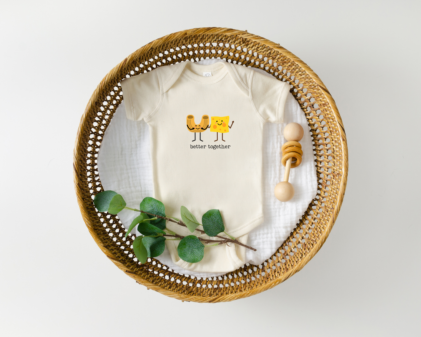 A short sleeve cotton baby onesie with a cute graphic of a macaroni noodle and cheese with the phrase "better together" styled in a basked with a baby toy.