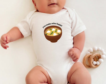 A baby wearing a short sleeve cotton baby onesie with a graphic of miso soup and the playful phrase "you make miso happy".