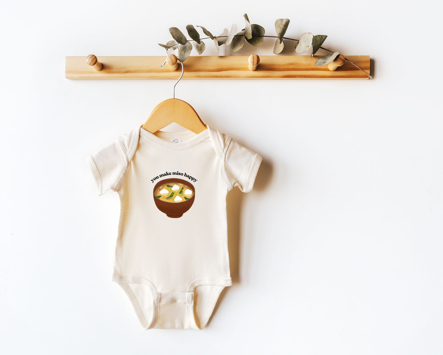 Short sleeve cotton baby onesie with a graphic of miso soup and the playful phrase "you make miso happy" displayed on a wooden hanger.