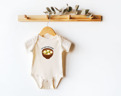 Short sleeve cotton baby onesie with a graphic of miso soup and the playful phrase "you make miso happy" displayed on a wooden hanger.