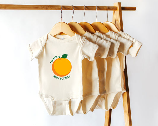 A short sleeve cotton baby onesie with the text 'Mama's Main Squeeze' and an orange graphic printed on the front, displayed among other bodysuits on a wooden hanger.