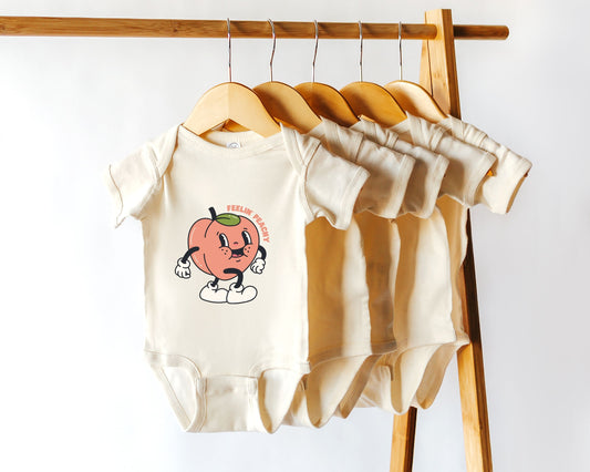 A short sleeve cotton baby onesie with a retro style graphic of a peach character with the text "feelin' peachy" displayed on a wooden hanger.