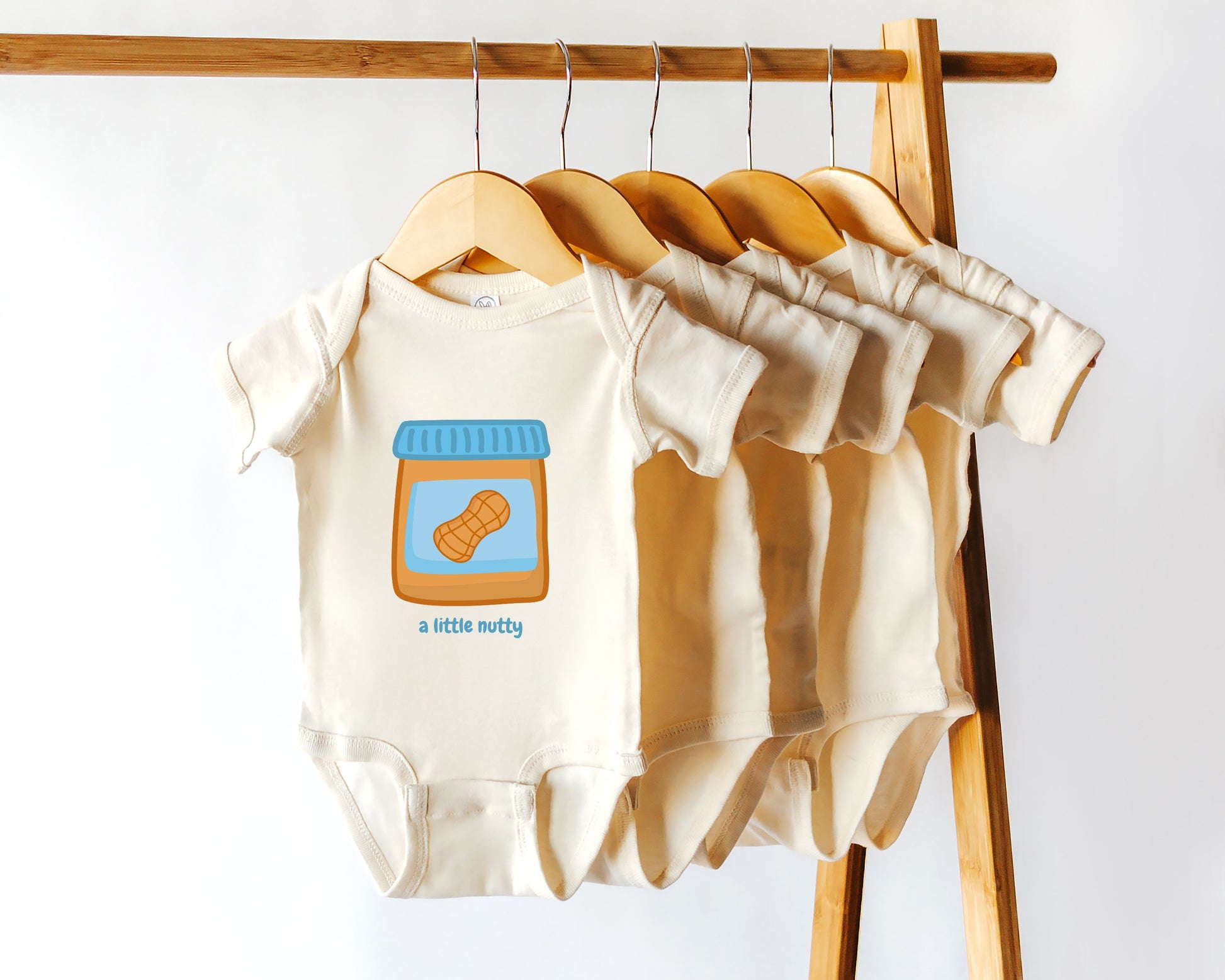 A series of cotton baby onesies with a graphic print of a jar of peanut butter and the phrase 'A little nutty'. The bodysuits are displayed on a wooden hanger rack.