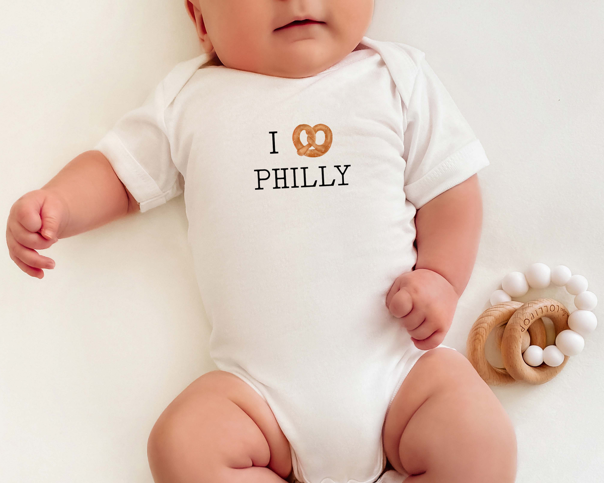 Baby wearing an short sleeve cotton bodysuit with a soft pretzel graphic for an I Heart Philly message.