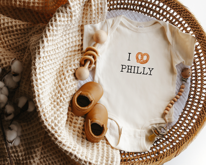 A baby bodysuit with 'I [pretzel] PHILLY' printed on the chest, styled in a wicker basket with a knit blanket.