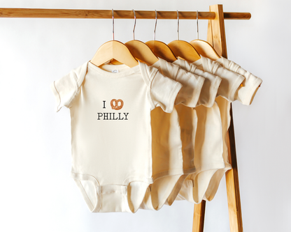 A set of cream-colored baby bodysuits hanging on wooden hangers from a wooden clothing rack. The front bodysuit features the text 'I' followed by a pretzel icon and 'PHILLY' in black.
