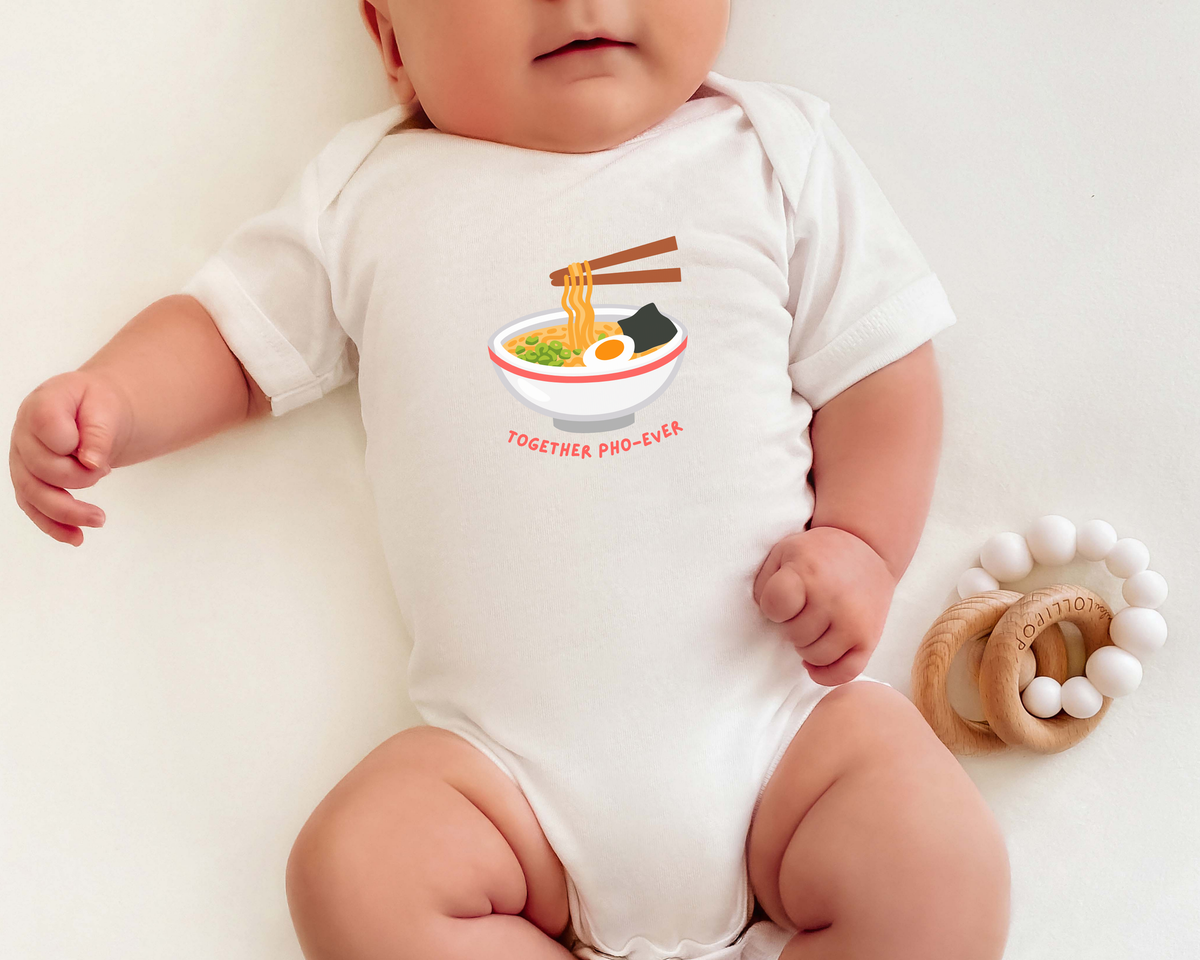 A baby wearing a white short-sleeve bodysuit with a graphic of pho and the text 'TOGETHER PHO EVER'. The baby is lying on a light cream surface next to a wooden teething ring.