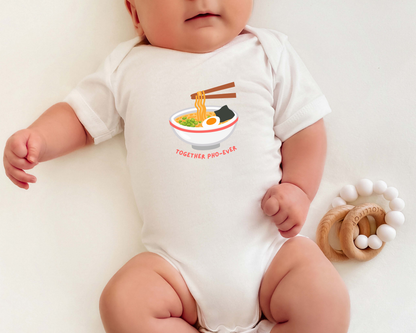 A baby wearing a white short-sleeve bodysuit with a graphic of pho and the text 'TOGETHER PHO EVER'. The baby is lying on a light cream surface next to a wooden teething ring.
