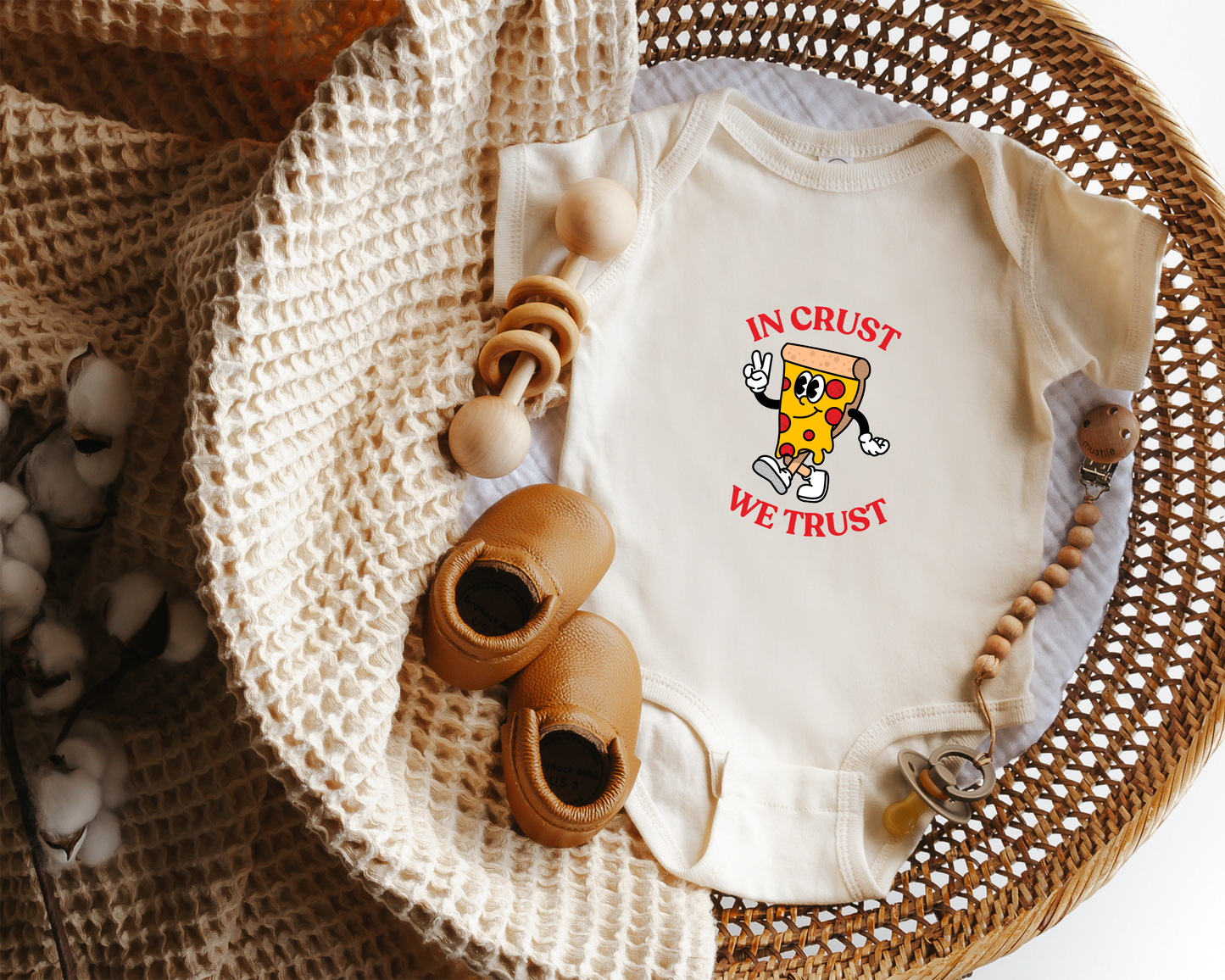 A baby onesie laid flat in a woven basket. The onesie is cream with a cartoon pizza character and the text 'IN CRUST WE TRUST' in red.