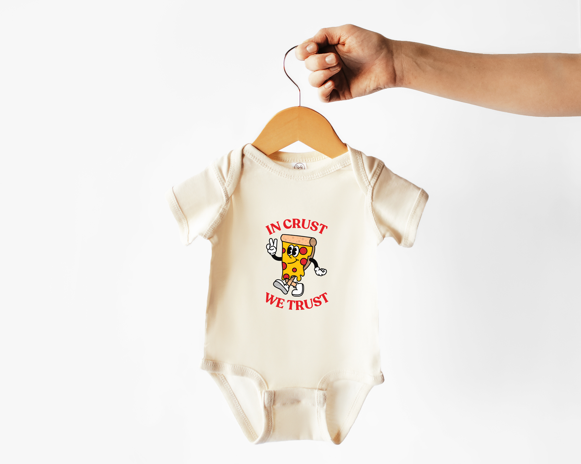 A cream-colored baby bodysuit with a cartoon pizza graphic and the text 'IN CRUST WE TRUST' in red, hanging from a wooden hanger held by a person's hand against a white background.