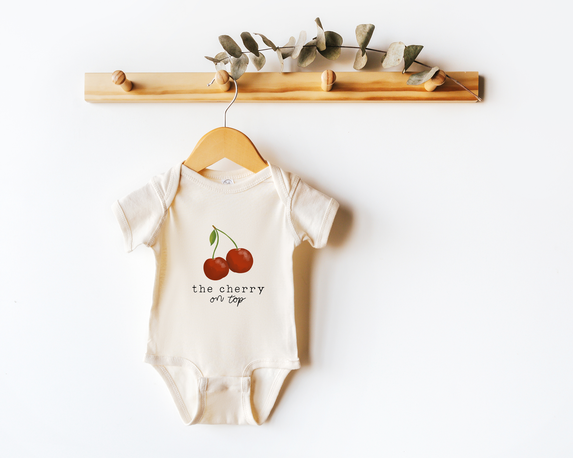 Baby onesie with cherry graphic and the text "the cherry on top" shown on a hanger.