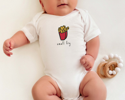 A baby wearing a white short-sleeve bodysuit with a small french fries graphic and the text 'small fry' on the chest, lying on a white background next to a wooden teething ring.