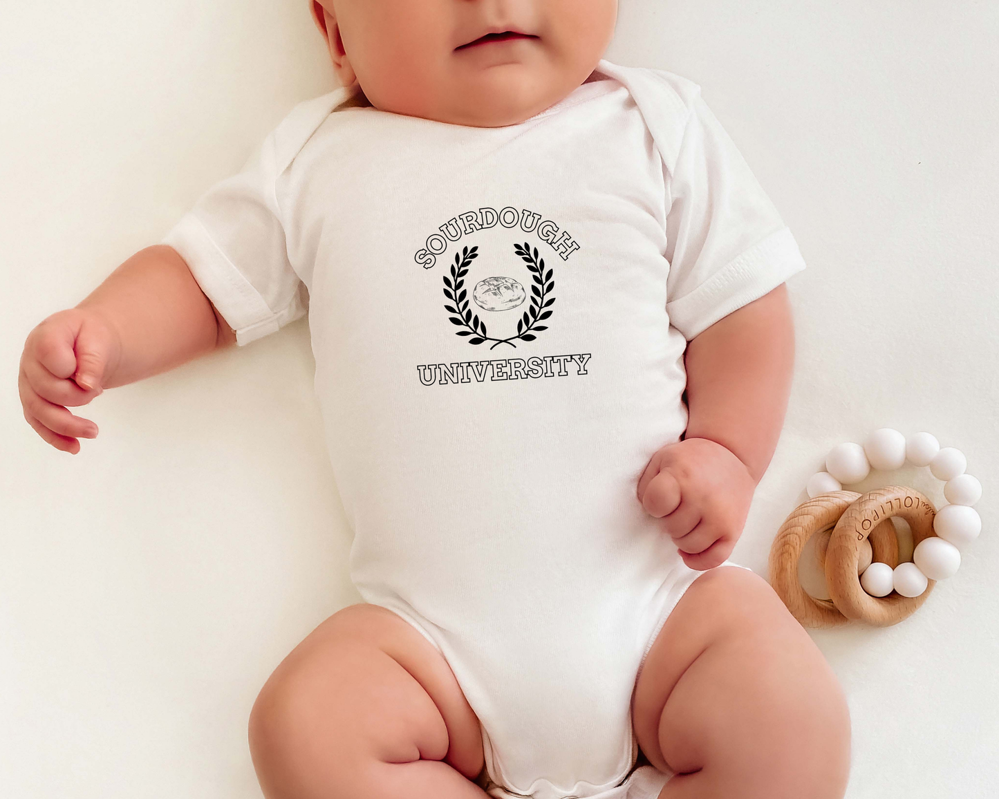 A baby wearing a white short-sleeve bodysuit with a black 'Sourdough University' graphic on the chest, lying on a white background next to a wooden teething ring toy.