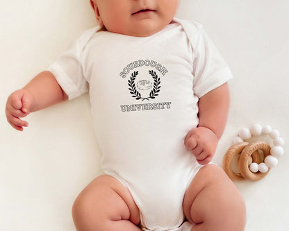 A baby wearing a white short-sleeve bodysuit with a black 'Sourdough University' graphic on the chest, lying on a white background next to a wooden teething ring toy.