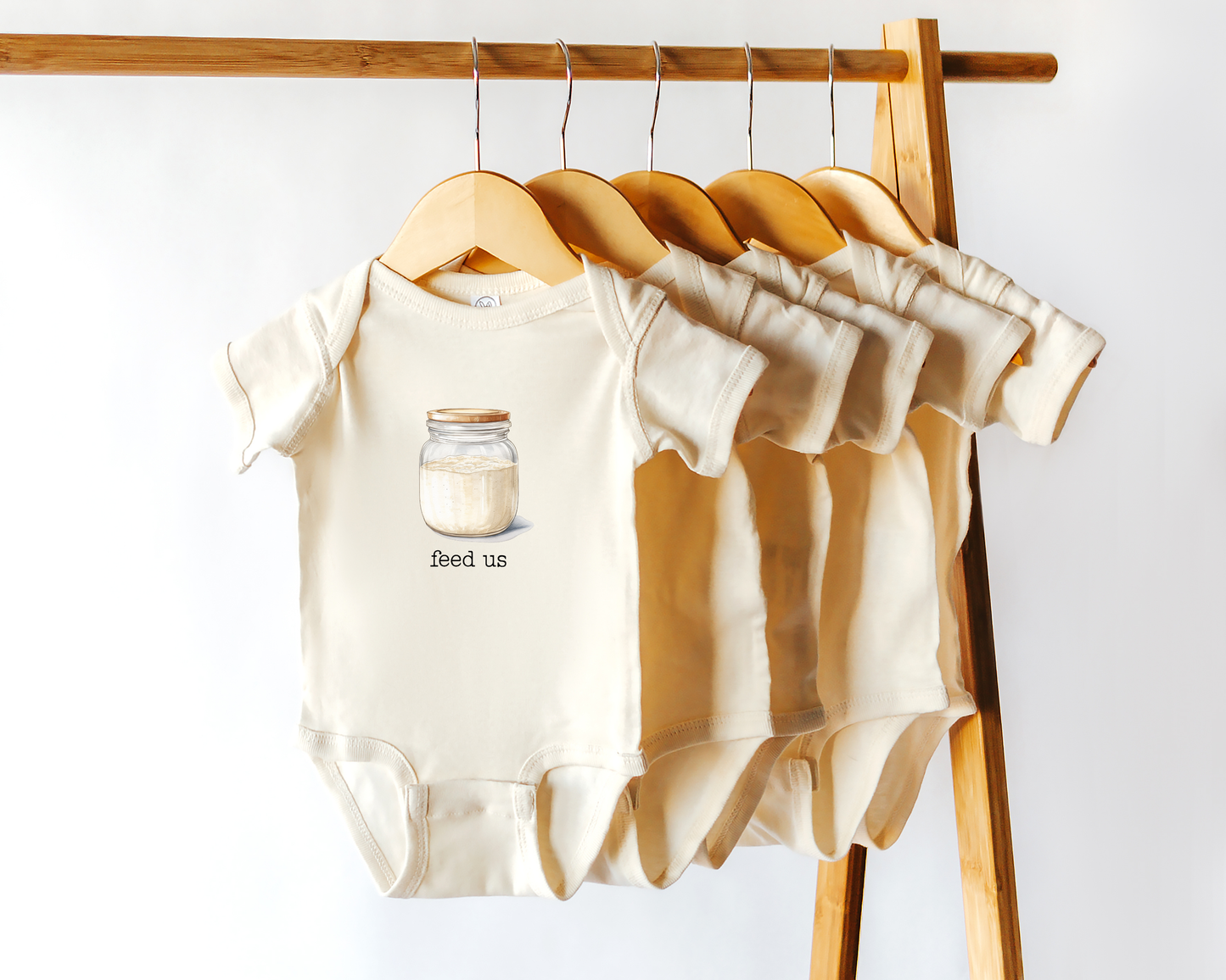 Short sleeve cotton baby onesie with a sourdough starter jar graphic and the text "feed us" displayed on a wooden clothing rack. 