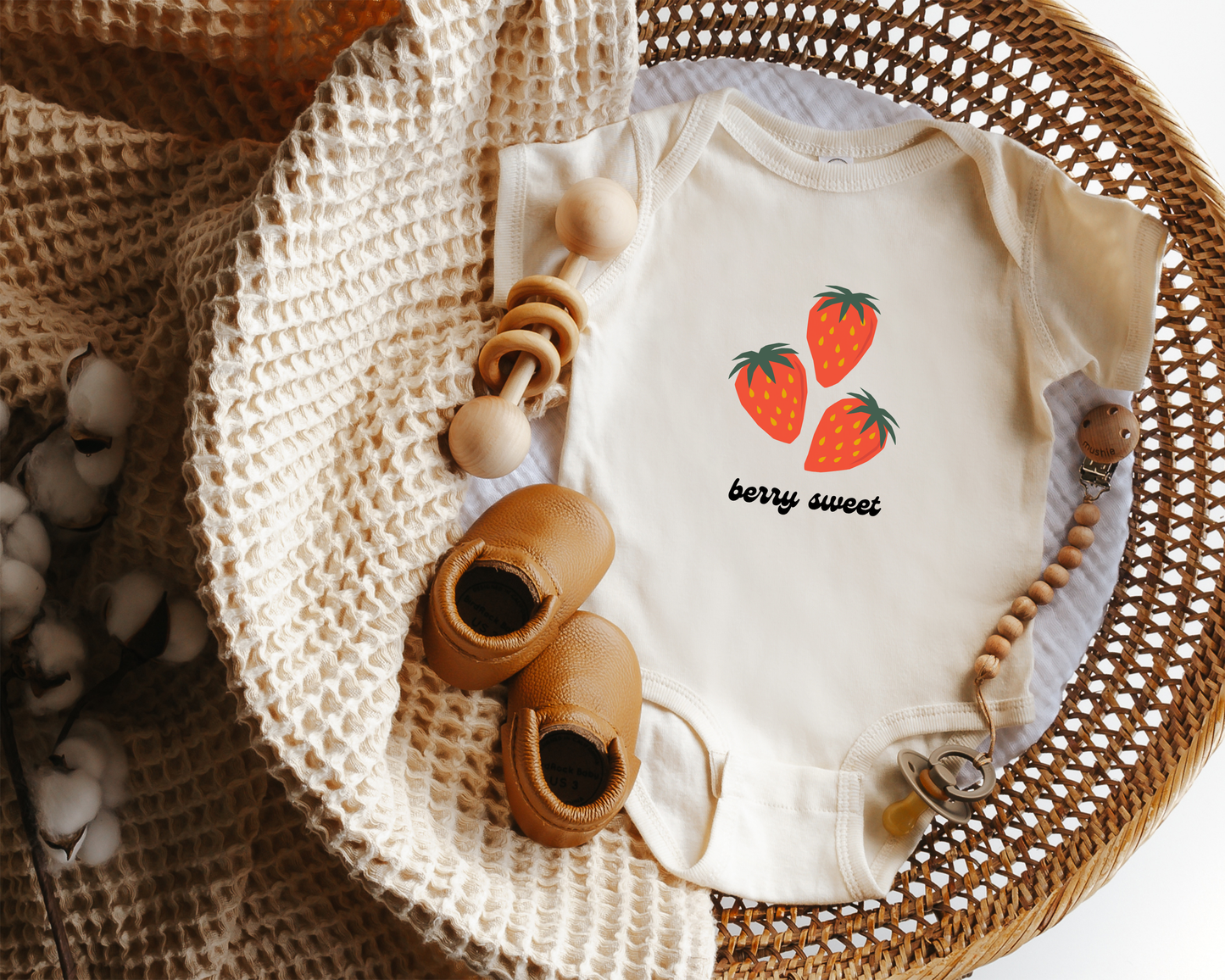 A short sleeve cotton baby onesie with red strawberry graphics and the text "berry sweet" displayed in a wicker basket.