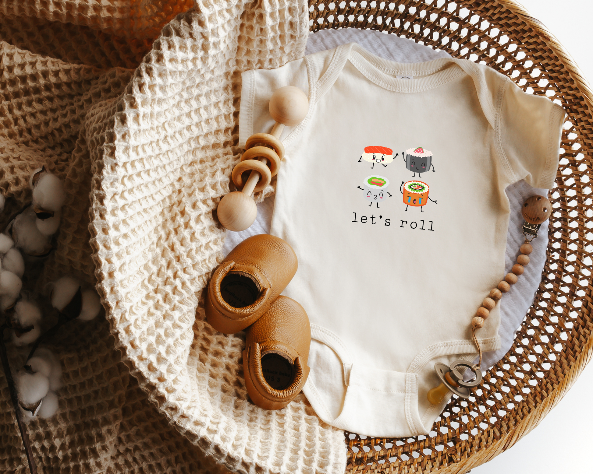 A short sleeve cotton baby onesie featuring graphics of sushi and the text "let's roll". Styled in a basket with a baby blanket, toy and baby shoes.