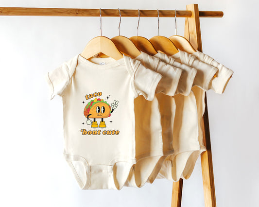 A cotton baby onesie with a retro taco graphic and the text 'Taco 'Bout Cute' hanging on a wooden rack among other similar bodysuits.