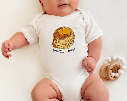 A baby wearing a white short-sleeve bodysuit with a waffle print and the text 'waffley cute' next to a wooden teether on a light cream background.