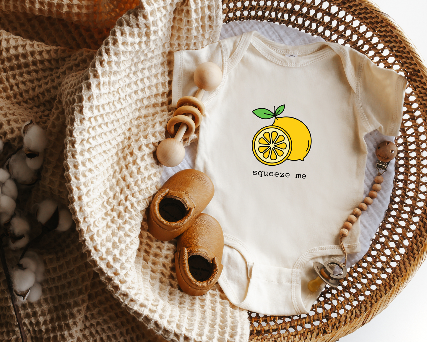 A short sleeve cotton baby onesie with a yellow lemon graphic and the text "squeeze me" displayed in a wicker basket.